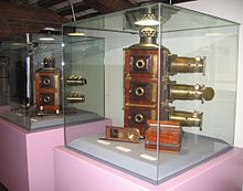 Two glass cabinets containing magic lanterns. On the left a bi-unial magic lantern by W.Tyler is displayed. The right-hand cabinet contains the J. H. Steward tri-unial mahogany magic lanterns with brass lenses.