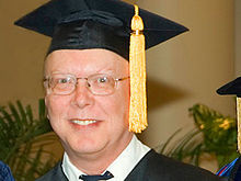 Profile shot of a bespectacled man in his early fifties, wearing a mortarboard with a yellow tassel