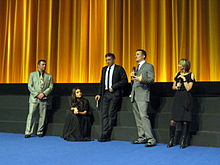 Scott Franklin, Mila Kunis, Vincent Cassel, Darren Aronofsky, and a moderator stand on a stage with a golden curtain backdrop wearing formal attire and discussing Black Swan