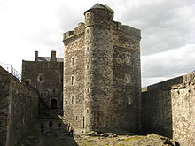 View of castle within its courtyard with a stone building over three stories tall in the middle.  The building is flanked by stone walls on both sides that are half as tall.
