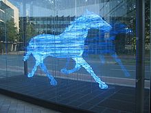 Photograph of a sculpture of a trotting horse. The sculpture was made using a series of horizontal bands. Each band is made from a glass tube that glows blue. The bands are stacked to indicate the form of a horse; the separation between the bands has about the same width as the glass tubing. The sculpture is inside a large glass box that has been placed on the plaza between some office buildings.