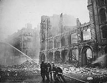 Historical photograph of a building severely damaged by air-raid bombing; firefighters are putting out a blaze in the ruins.