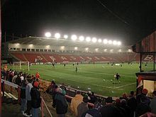 A football stadium at night. In the foreground is the pitch, behind it is a grandstand with floodlights and orange seating.
