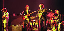 Dylan together with three musicians from The Band onstage. Dylan is third from left, wearing a black jacket and pants. He is singing and playing an electric guitar.