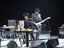 Dylan, dressed in a black western outfit with red highlights, stands onstage and plays the keyboards. He gazes to the left of the photo. Behind him is a guitar player, dressed in black.