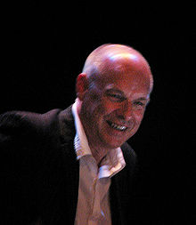 Brian Eno—middle-aged Caucasian man with a shaved head—smiles while wearing a black blazer and white shirt.