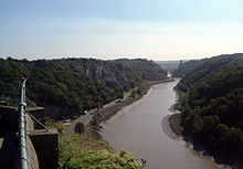 River flowing though a steep sided valley. In the distance is a suspension bridge supported by towers. In the left foreground is a handrail.