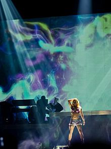 Image of a blond female performer. A big screen is placed behind her. The screen is filled with cosmic and psychodelic images.