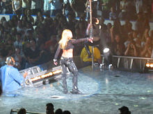 Image of a blond female performer. She is turning her back to the camera, near the end of the stage. She is wearing black pants and a black top with long sleeves, showing her midriff. In front of her, a female dancer is climbing into a pole. A crowd of people clapping, screaming and taking pictures stands can be seen in the audience.