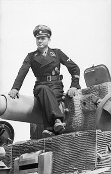 A man, wearing dress uniform and a cap, sits on top of a tank barrel; the tank is not fully in view.