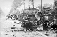 Several destroyed vehicles line the side of a tree and hedge lined road. A destroyed gun, twisted metal and debris occupy the foreground.
