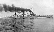 A large warship plows through the water, thick black smoke pouring from its two central smoke stacks.