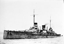 A large, light gray warship sits in harbor, the two forward gun barrels are turned slightly to the left.