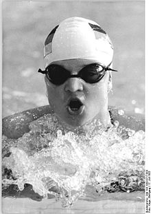  A female swimmer wearing dark goggles and a white cap with the flag of East Germany on it. Her head is above the water and she is taking a breath with her mouth open as her arms push the water to the side during the breaststroke.