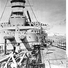 Two long gun barrels jut out of a wide, round gun turret aboard a warship; the deck is covered in heavy chains, winches, and cranes