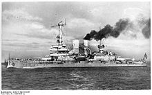 A large warship, thick black smoke pouring out of its rear funnel, steams through the calm sea
