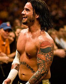 A side-view of a topless, caucasian male with shoulder length black hair. Several tattoos are visible on his arms and stomach, and he has white tape around both his wrists and his right elbow.