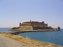 A large ruinous castle with concentric walls and towers located on an island that is connected to the shore by a causeway