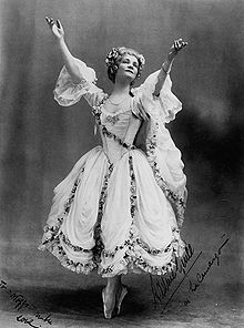 A woman in a white ballet skirt stands on the tips of her toes, arms extended upwards