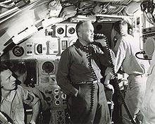 Submarine commanding officer using a microphone to address the ship's crew over the public address system in the submarine's control room with another officer facing away in background.