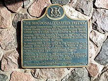 A blue plaque on a stone wall. The plaque has a yellow border, and is mostly rectangular in shape, with the long end oriented horizontally. However, the top side has a camel hump in the centre, with a circle centred at the top of the hump. Inside the circle is an Ontario coat-of-arms. The plaque reads: THE MACDONALD CARTIER FREEWAY This plaque commemorates the completion of the Macdonald-Cartier Freeway (Highway 401), the longest freeway operated without tolls by a single highway authority in North America. Covering 510 miles between Windsor on the United States border and the Ontario-Quebec boundary, it serves the richest economic region in Canada. In January, 1965, it was named by The Honourable John P. Robarts, Prime Minister of Ontario, in honour of the two founding architects of the Confederation of Canada, Sir John A. Macdonald and Sir Georges Etienne Cartier. This site is located on the last section of construction, consisting of 15 miles between Ivy Lea and Highway 2, which was completed on October 11, 1968.