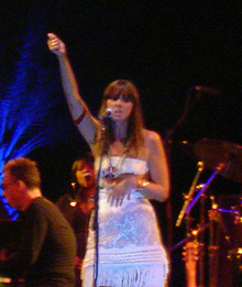 A female vocalist wearing a white dress performing on stage. Some members of a string section present in the left-hand side and part of a drum kit present on the right