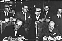 A black and white photograph of two mature and formally dressed men seated in chairs, signing some papers over a large table while a group of six men in suits stand behind their seats.