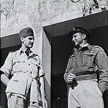 Two men in military uniform standing outside of a buildind talking to each other. The man on the left is wearing a lighter coloured uniform with short sleaves. The man on the right is wearing a long sleaved, dark jacket.
