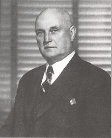An unsmiling balding white man in a three piece suit