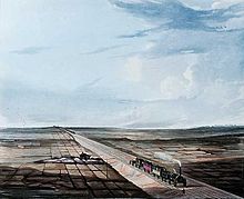A very early steam locomotive pulling four open carriages under a cloudy blue sky along a track slightly built up from the surrounding flat countryside. The train has just passed a small farmhouse and is approaching a gentleman who is standing by the side of the track.