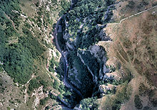 Looking down into the gorge with a road with cars on it running from the top to the bottom of the picture. To the right are exposed rocky cliffs with trees on some ledges. To the left are less steep slopes covered in vegetation.
