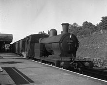Large dark steam locomotive at a railway station platform, with a steep earth bank behind it