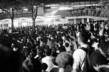 B/W image of hundreds of people orderly sitting outdoors, with an unfurled banner