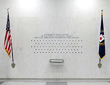 A white wall featuring an inscription, several dozen stars placed in five rows, a book, framed by the U.S. flag on the left and a flag bearing the CIA's seal on the right.