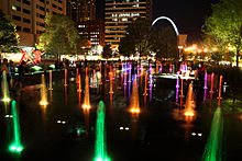 In this low aerial night shot, numerous fountains shoot water into the air, illuminated by colored lights.