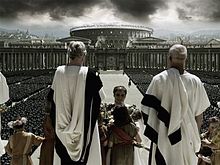 Several men in white robes are facing away from the image, at the top of large steps. A man is at the center of the image being handed flowers by a girl. In the background are rows of thousands of soldiers and members of a large crowd. In the distance, the Colosseum can be seen along with other buildings in Rome. Dark clouds are visible in the sky.