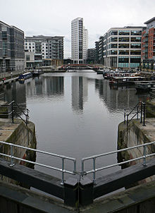 A night-time scene of a dock containing a number of moored canal-boats to left and right and railings around the edges. At the front is part of a lock gate and steps leading down to the water. Around most of the dock are multi-storey modern buildings, some with lighted ground-floors and seats and decorative objects outside. The most prominent of these, at the far end, is a twenty-storey building with curving facades.