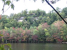 A cliff with a small shelter at its top, viewed from across a lake. The leaves in the trees growing from the slopes are green, pink, purple, and red.