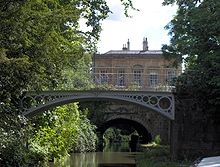 A iron bridge spanning water. In the background is a yellow stone building. On the left tress reach out over the water.