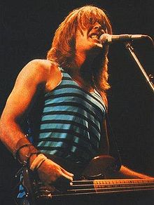 Thirty-one year old Williams is at a microphone with teeth clenched, he strums his right hand on the bass while the left is out of view. His red-brown hair is over collar length. He wears a singlet and has a leather arm band.