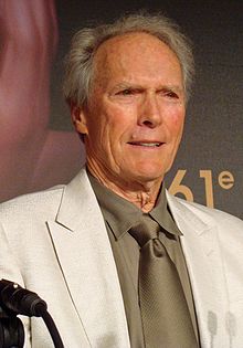 An older man is at the center of the image smiling and looking off to the right of the image. He is wearing a white jacket, and a tan shirt and tie. The number 61 can be seen behind him on a background wall.