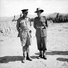 Two officers in tropical military uniforms standing on sandy ground, a larger body of troops is visible in the right background in front of stunted trees and more distant hills