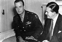 A monochrome photograph of two men, one about 30 years old wearing a military pilot's uniform of the rank colonel, seated, leaning forward to the left, looking left, and the other in his mid-40s wearing a suit jacket, white shirt and tie, with dark hair oiled, parted and combed
