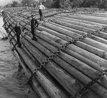 Three men in work clothes stand on an enormous raft of logs held together with cable chains. In the background, another three men work on a distant part of the raft, only part of which is visible. The pile of logs appears to be taller than any of the men.