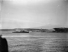 A black and white photograph showing a warship firing its armament at positions on the foreshore, while in the distance landing craft move towards the beach