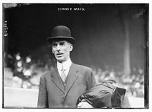A man standing in a baseball stadium wearing a suit and bowler hat. He has a coat folded over his left arm.