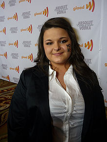 18-year-old Caucasoid female with a left-nostril piercing and the letters "NO H8" painted on her left cheek is wearing a white shirt and black coat and standing in front of a banner advertising the "GLAAD awards" and "Absolut Vodka".
