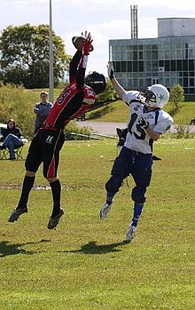 Two football players, an offensive receiver and a defensive cornerback, both reach for a thrown football. The cornerback is in front of the receiver with the ball almost secured in his hands.