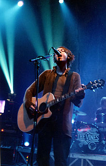  Nicholls is shown singing into a microphone while strumming a guitar.