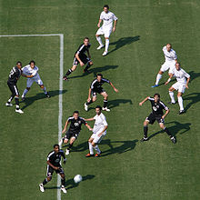 Six soccer players in black and five in white views from above look up for a moving soccer ball coming toward them.